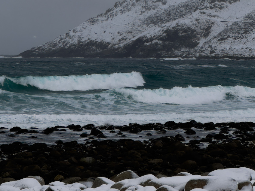 Hohe Wellen an einem mit großen, dunklen Steinen bedecktem Strand, die zum Teil von Schnee bedeckt sind. Im Hintergrund ein hoher schneebedeckter Berg.